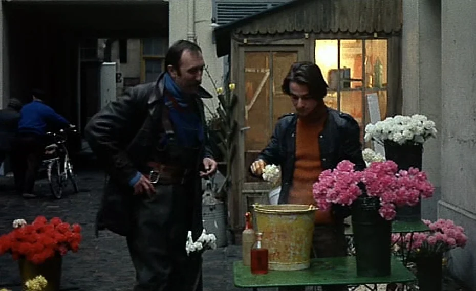 Bed & Board (1970) movie still: medium — A small outdoor floral stand is situated in a paved urban courtyard area. A middle-aged man in leather…