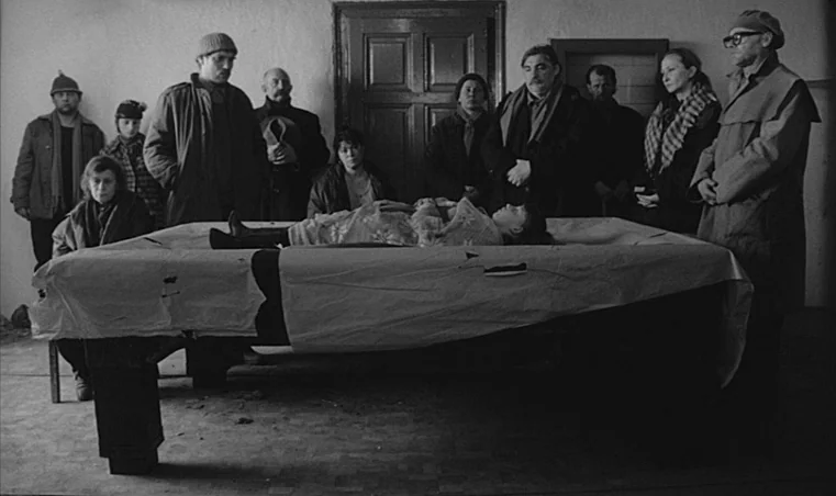 Sátántangó (1994) movie still: wide — A bare room with white walls and a large, central table covered in paper. A group of adults stands around the…