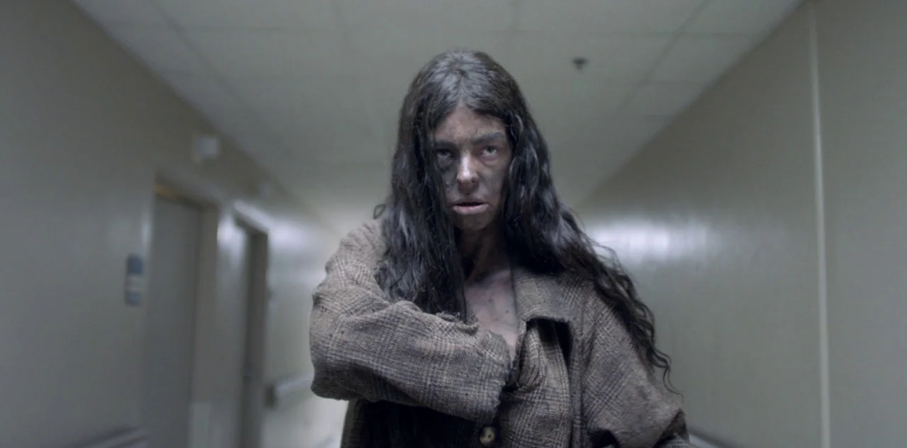 Darlin’ (2019) movie still: medium — A long, sterile hospital corridor with pale walls and closed room doors. A woman with disheveled hair and a…