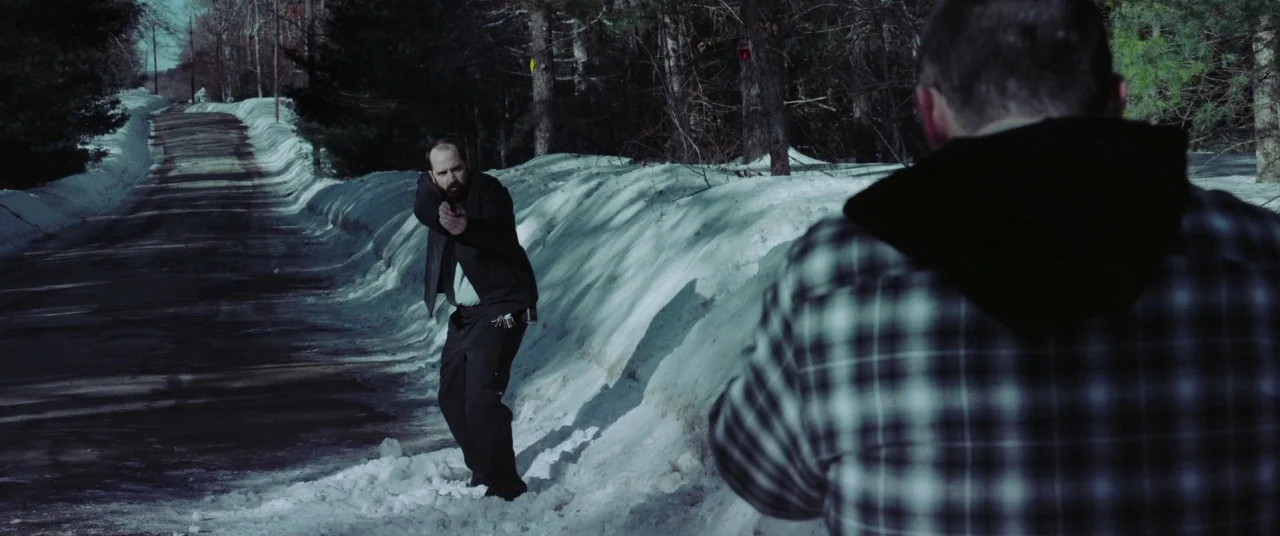 The Mind’s Eye (2015) movie still: medium — A snow-covered road is flanked by dense woods and high banks of shoveled snow. One man in a blazer aims…