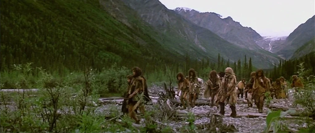 Clan of the Cave Bear (1986) movie still: wide — A wide valley floor features a dry, rocky riverbed flanked by dense evergreen forests. A group of…