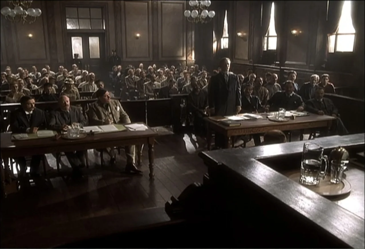 Vendetta (2022) movie still: wide — A wood-paneled courtroom filled with people sitting at long desks during the day. A man stands in the center…