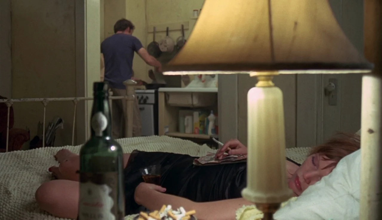 Fat City (1972) movie still: medium — A cluttered apartment shows a woman resting on a bed in the foreground. In the blurred background, a man…