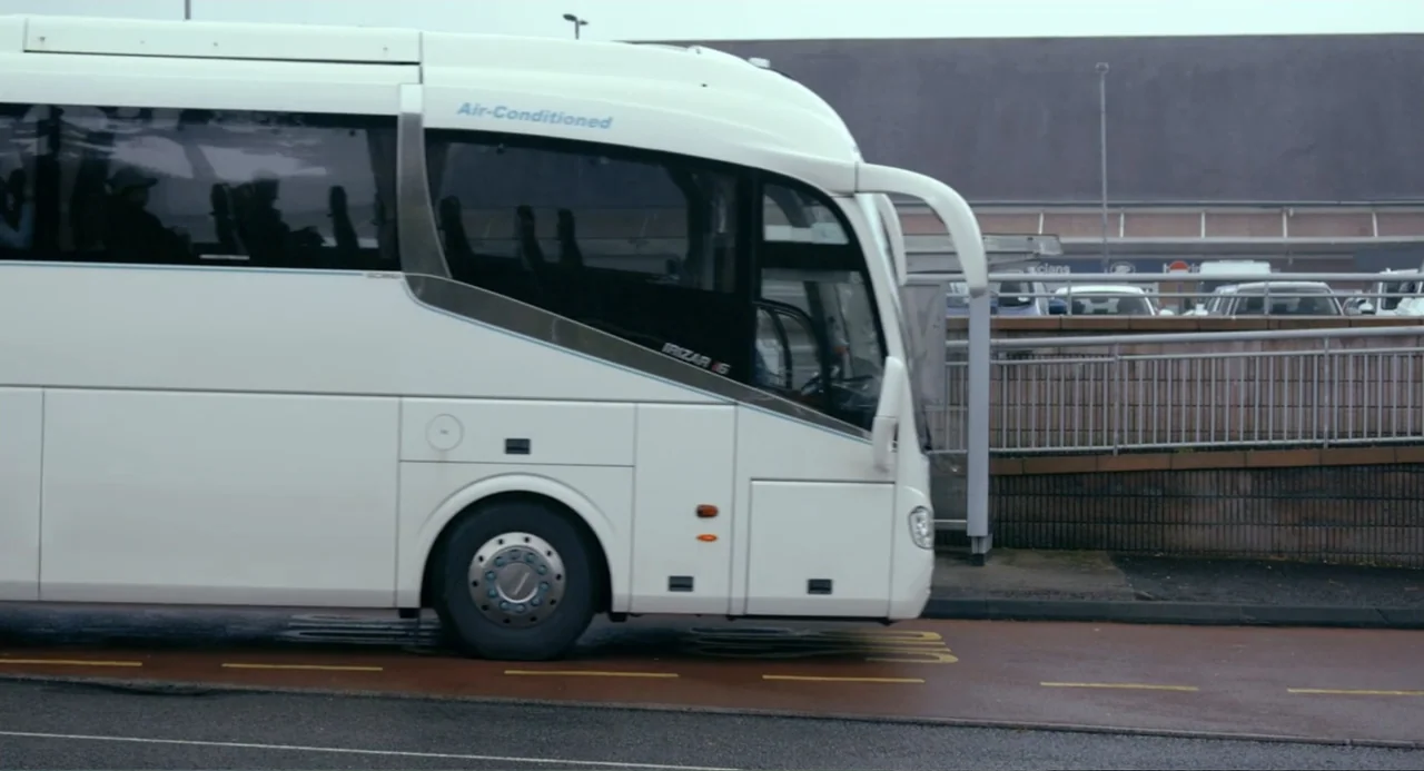 Living Out Loud (1998) movie still: medium — A white coach bus drives along a road next to a metal pedestrian barrier. The background consists of a…