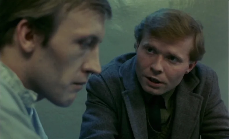 No End (1985) movie still: medium — An austere room with flat-painted green walls and minimal decoration provides a stark backdrop. Two men engage…