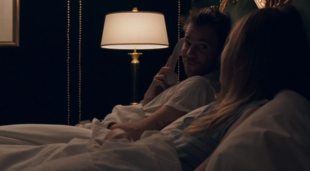 Somewhere (2010) movie still: medium — A dark hotel room features a single lit bedside lamp. A man talks on the phone while looking at a woman in…