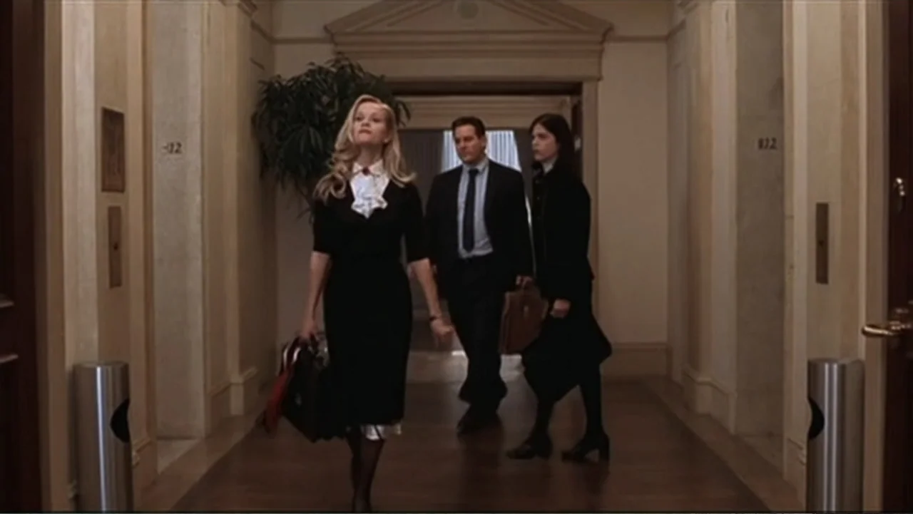 Introducing, Selma Blair (2021) movie still: fullbody — A well-lit hotel corridor features wooden floors and neutral beige walls. Three people in…