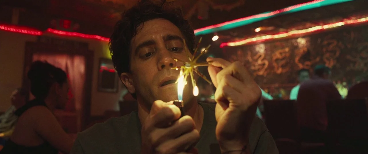 Stronger (2017) movie still: closeup — A dimly lit bar space with red and green neon lighting elements visible. A man looks down at a metal object…