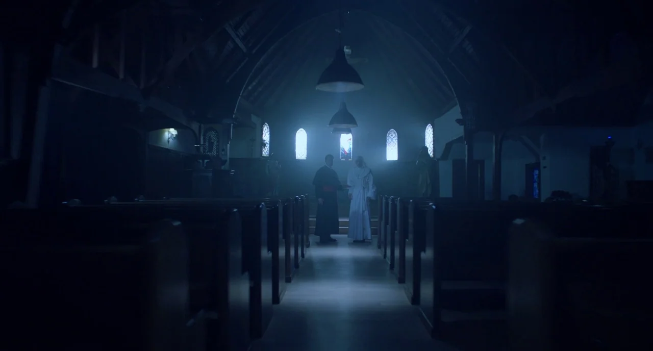 Nightmare Cinema movie still: wide — A dark church interior with long wooden pews lining a stone center aisle. Two figures in clerical attire stand…