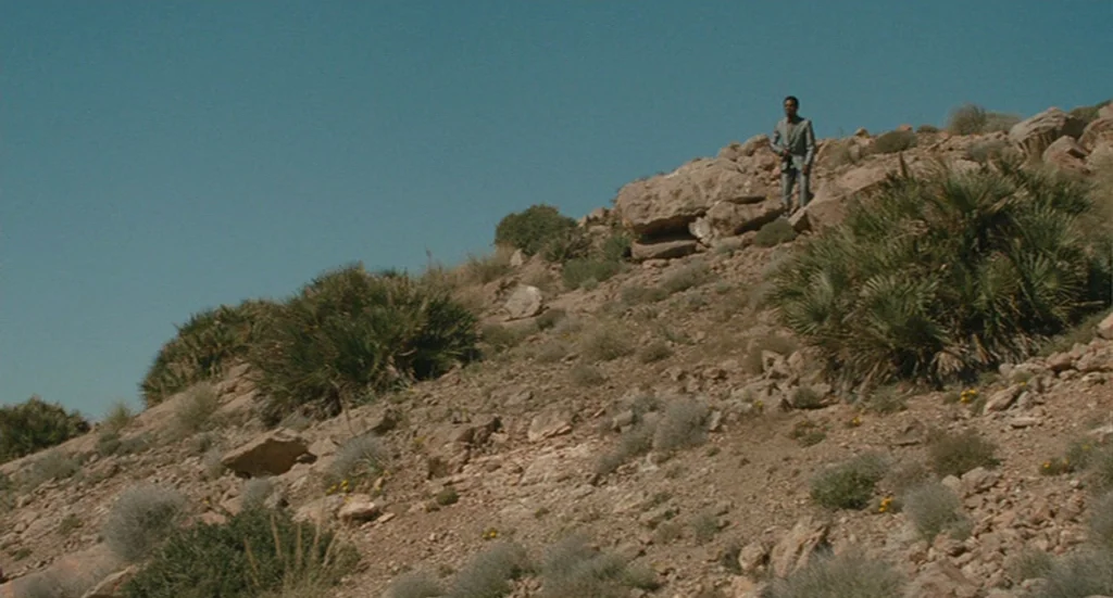 The Limits of Control (2009) movie still: wide — An arid, rocky hillside features sparse desert vegetation and scattered large boulders. A man in a…