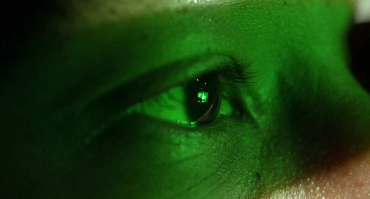 Femme Fatale (2002) movie still: closeup — A person's eye is illuminated by a sharp, green light source in darkness. The reflection of a small…
