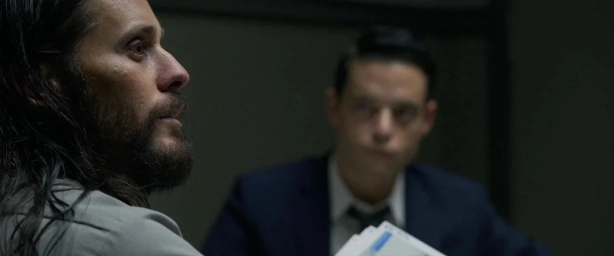 The Little Things (2021) movie still: medium — A dimly lit room with plain walls and overhead fluorescent lighting. Two men face each other while…