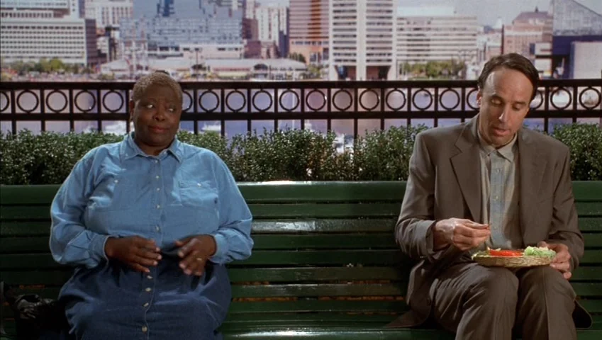Making of: Castle Freak (1995) movie still: medium — A green wooden bench sits in front of a metal railing overlooking a cityscape. A woman and a…
