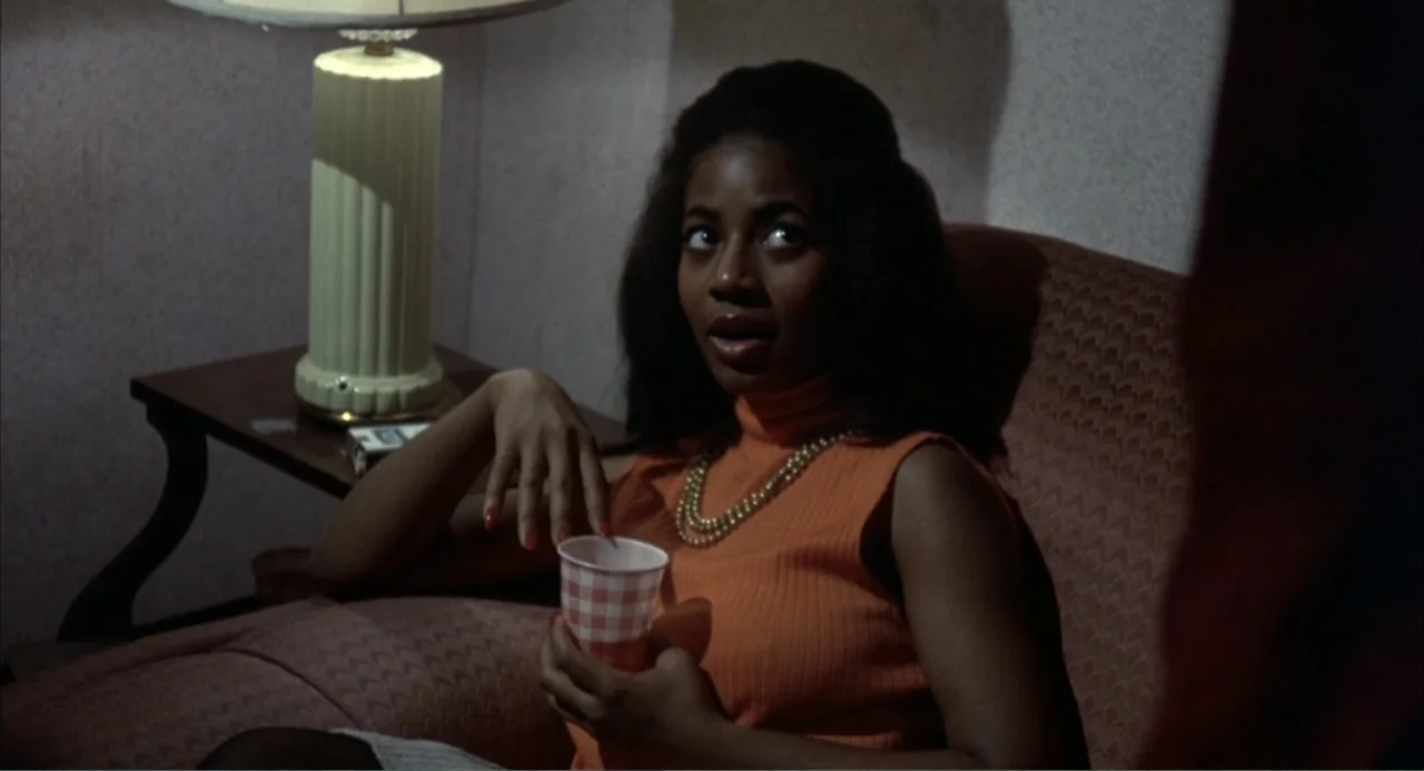 Cooley High (1975) movie still: medium — A woman sits in a chair in a 1970s living room with wallpaper walls. She holds a small paper cup while…