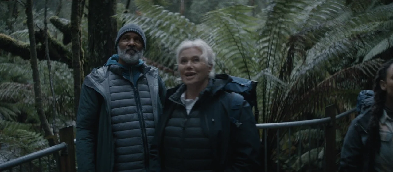 Force of Nature: The Dry 2 (2024) movie still: medium — A damp trail lined with large ferns and trees under an overcast sky. Two older adults in…