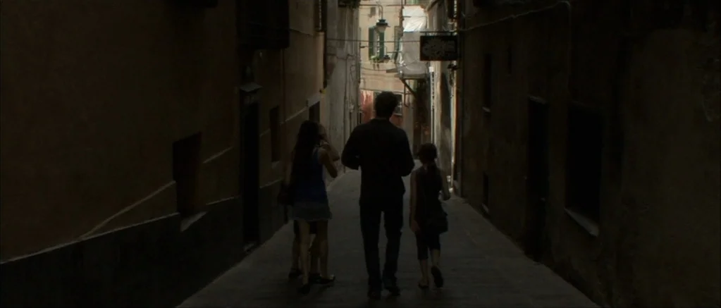 Genova (2008) movie still: fullbody — A narrow stone passageway between tall, shadowed buildings leads to an open area. Two adults and a child walk…