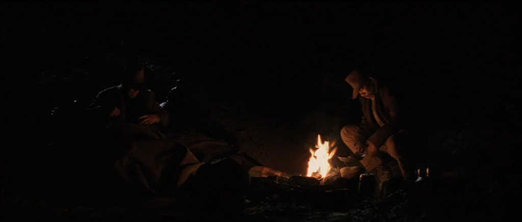 Unforgiven (1992) movie still: wide — Two men sit on the ground at night near a small, bright campfire. The rest of the campsite is enveloped in…