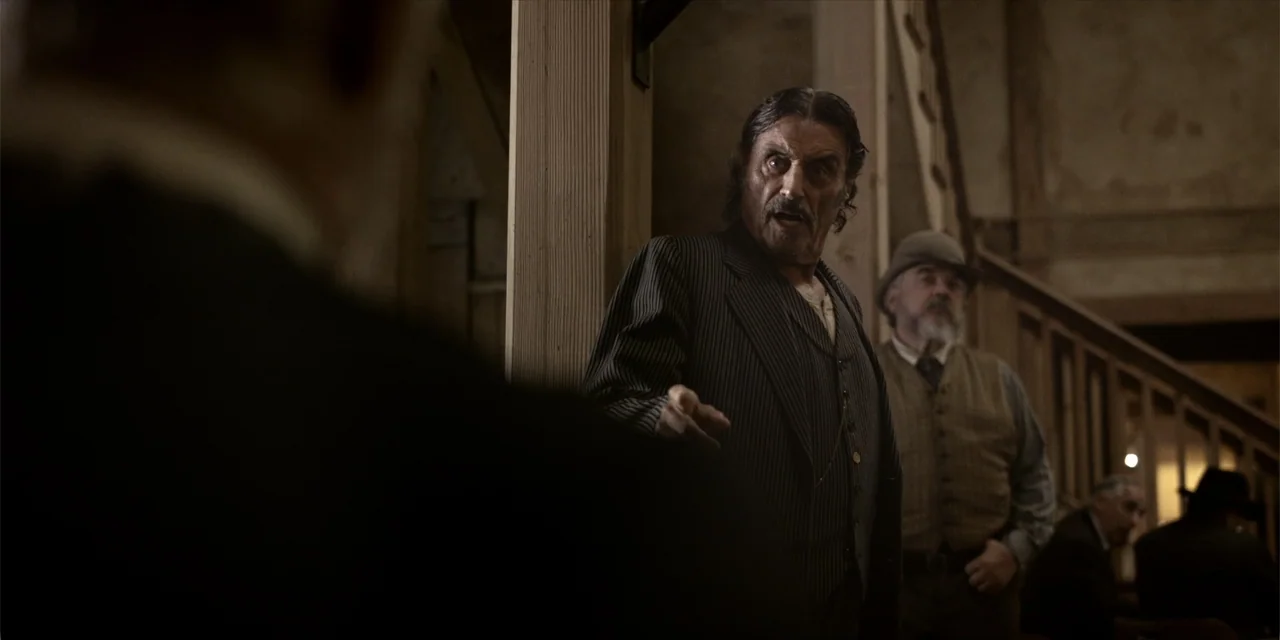 Deadwood: The Movie (2019) movie still: medium — A dimly lit 19th-century wooden saloon features a staircase and rough-hewn support columns. A man…