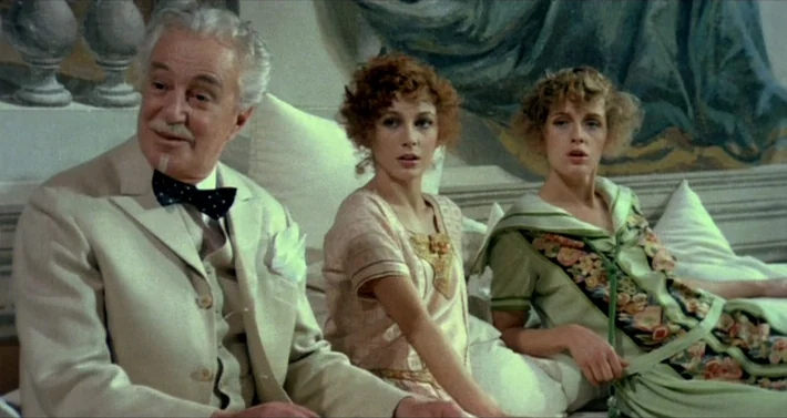 Blood For Dracula (1974) movie still: medium — A 1920s bedroom setting features ornate wall paintings and white bedding. An elderly man and two…
