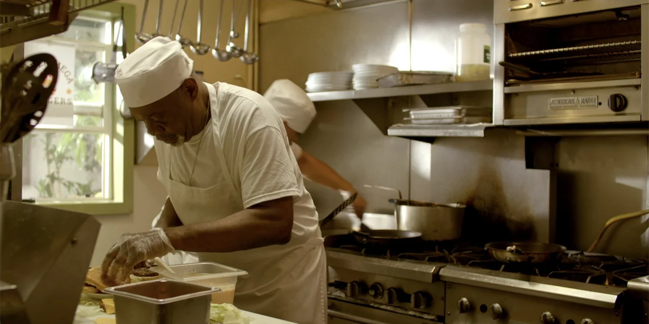 7th & Union (2021) movie still: medium — A commercial kitchen features stainless steel appliances, hanging metal cookware, and stacks of plates. A…