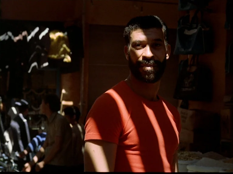 Fox and His Friends (1975) movie still: medium — A man stands in an outdoor marketplace with various textile goods hanging in the background. He is…