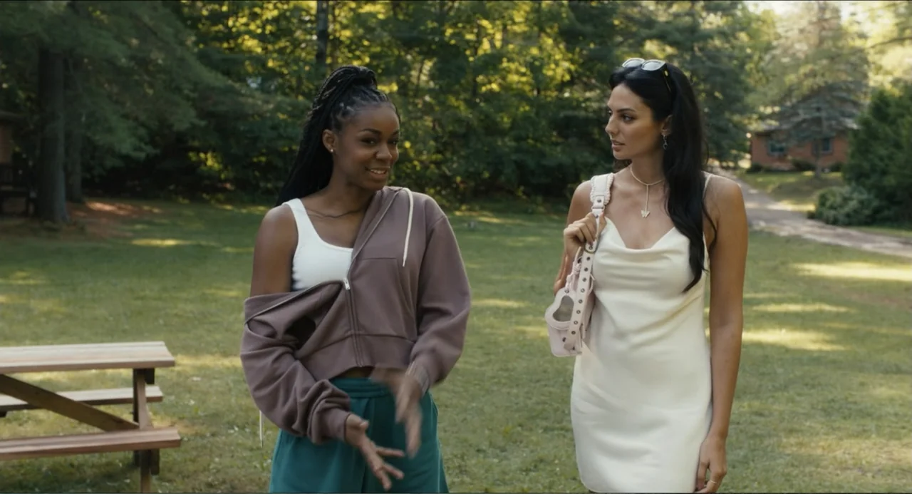 Hell of a Summer (2025) movie still: medium — A grassy clearing surrounded by dense woods features a wooden picnic table. Two young women walk…