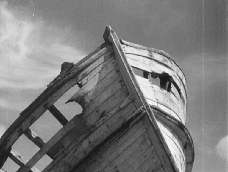 Redes (1936) movie still: closeup — A wooden boat hull sits in disrepair against a bright cloudy sky. The wood planks are splintered and decaying…
