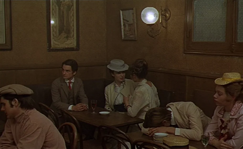 Two English Girls (1971) movie still: medium — A dimly lit room with brown textured walls decorated with framed prints and sconces. Several people…