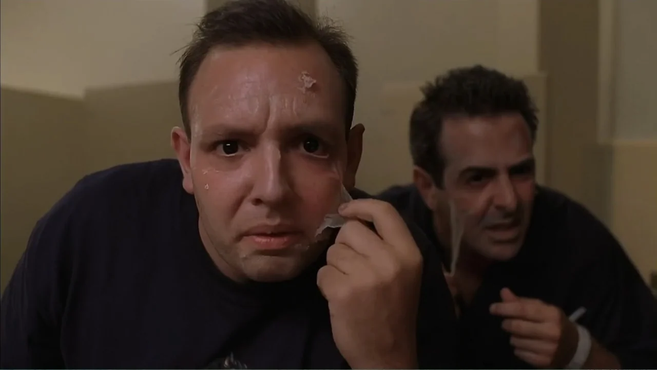 Day of the Dead 2: Contagium (2005) movie still: medium — A dimly lit room with plain walls features two men in navy shirts. The men peel adhesive…