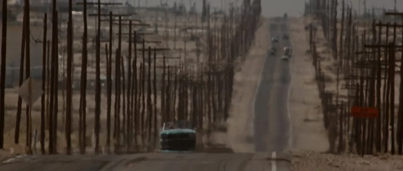 Thelma & Louise (1991) movie still: wide — A long, straight desert highway stretches toward the horizon under a hazy sky. Numerous wooden utility…