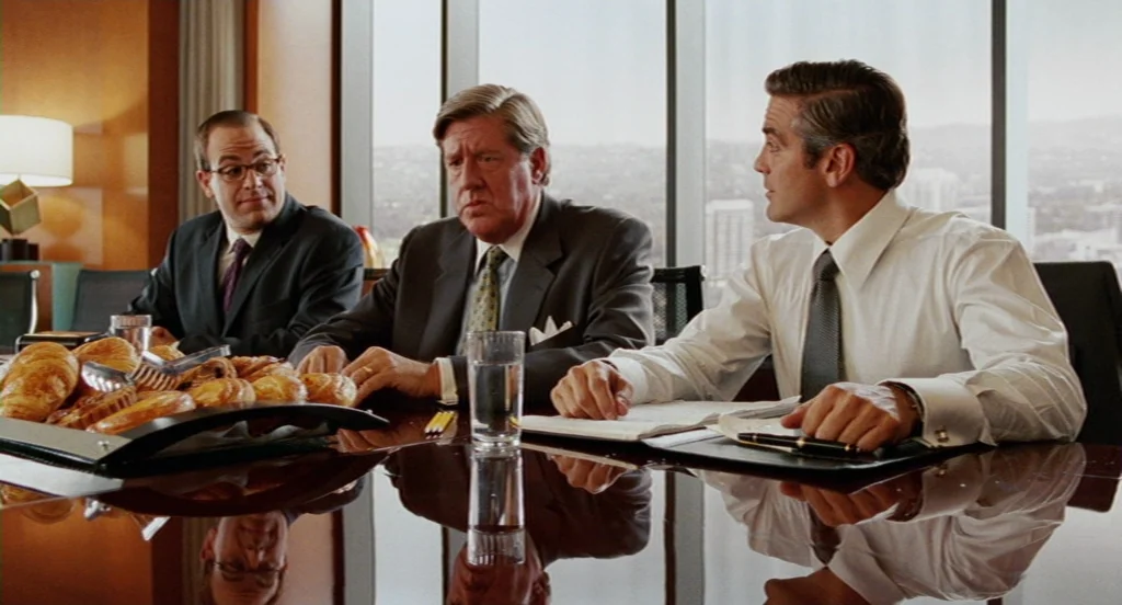 Intolerable Cruelty (2003) movie still: medium — A bright, modern conference room with floor-to-ceiling windows overlooking a city skyline. Three…