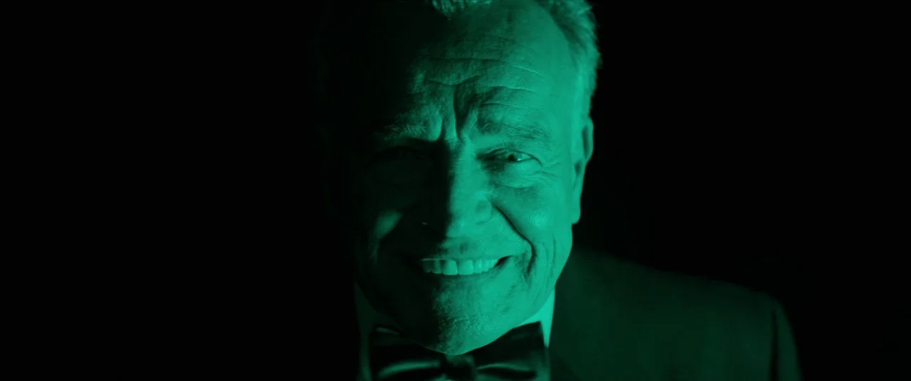 Tone Deaf (2019) movie still: closeup — An older man in a tuxedo stands against a completely black background. He is illuminated by a sharp green…
