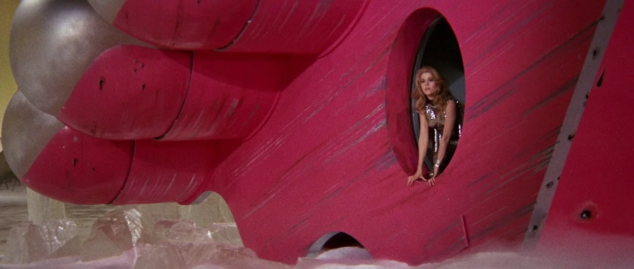 Barbarella (1968) movie still: wide — A woman looks out from a large pink, futuristic metallic vessel structure. She stands within a round portal on…