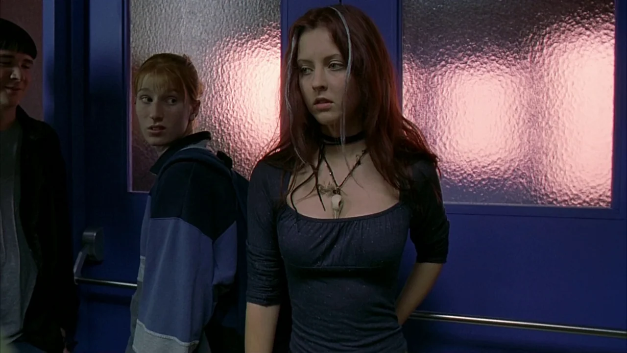 Ginger Snaps (2000) movie still: medium — A school corridor features blue metal double doors with translucent pink-tinted privacy glass panels. Two…