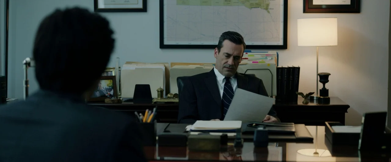 The Report (2019) movie still: medium — An office interior features a wooden desk, a wall map, and a desk lamp. A man in a suit sits behind the desk…
