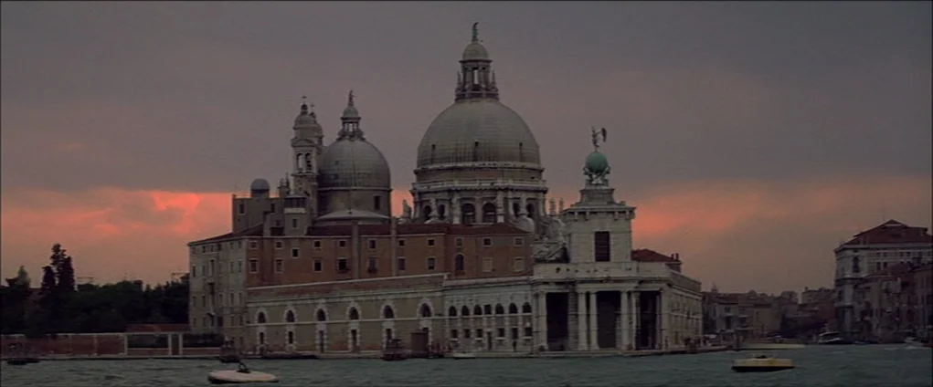 Death In Venice (1971) movie still: establishing — A historic church with large domes and stone walls sits at the water's edge. Dark, moody clouds…