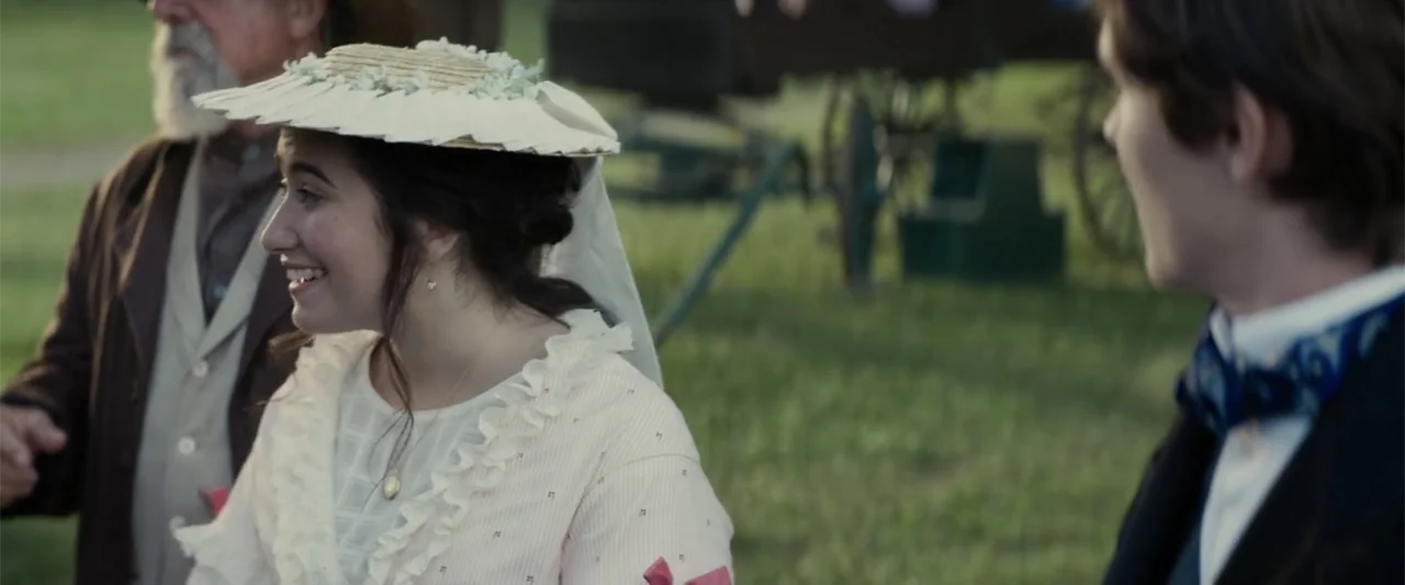 I Heard the Bells (2022) movie still: medium — A grassy outdoor area during the late afternoon sun with a wagon in back. A young woman in a summer…