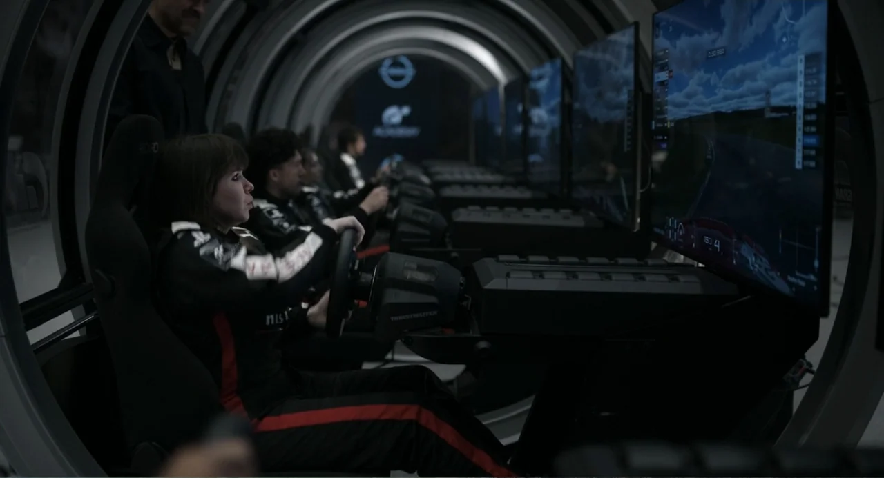 Gran Turismo (2023) movie still: medium — A long cylindrical room contains a row of professional racing simulator rigs. A young woman sits in the…