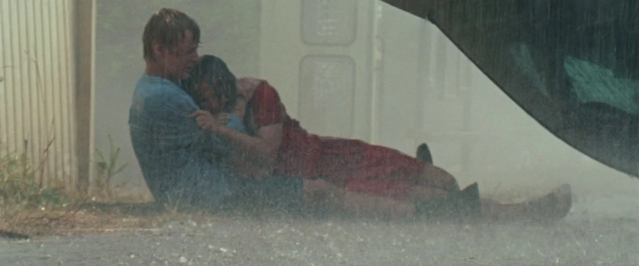 Adoration (2019) movie still: fullbody — A man and woman sit huddled together on wet pavement during a rainstorm. Rain splashes around them while…