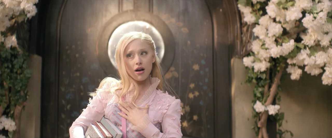 Wicked (1998) movie still: medium — A woman stands before a large arched wooden door adorned with painted floral motifs. She holds a stack of books…