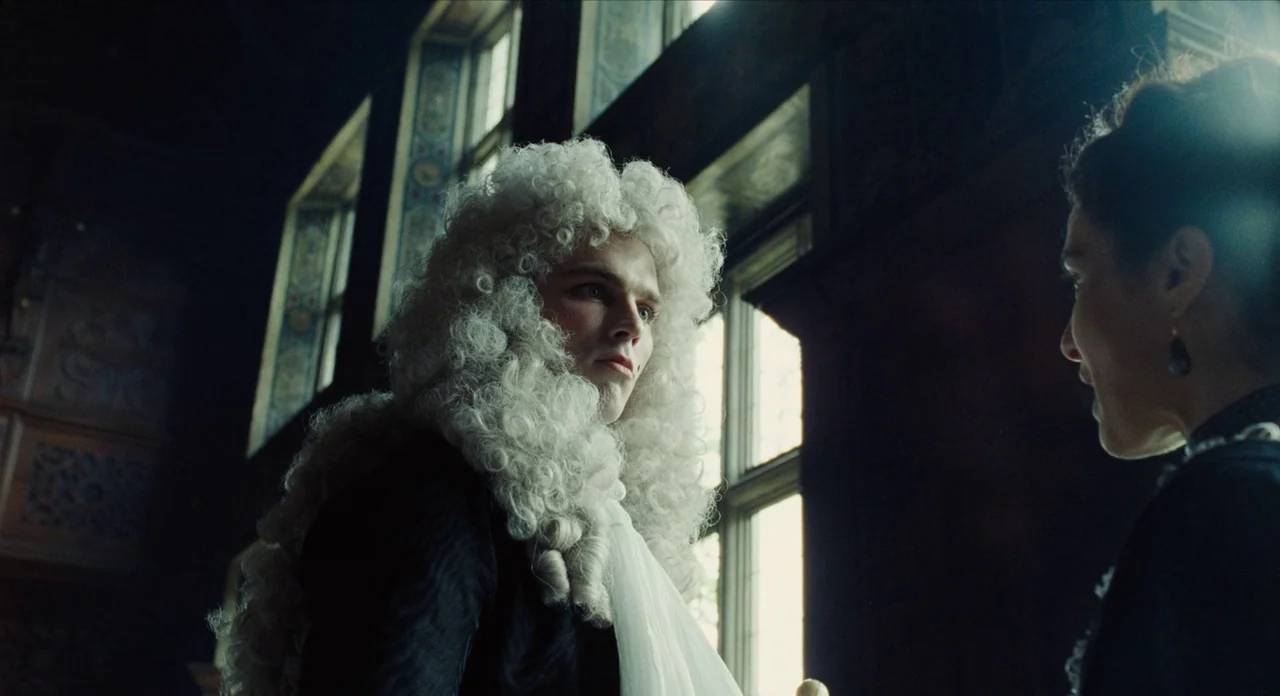 The Favourite (2018) movie still: medium — A dimly lit room in a historic manor features tall, multi-paned windows along the wall. A young man…