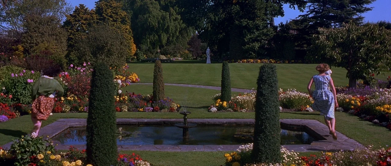 Heavenly Creatures (1994) movie still: wide — A well-maintained sprawling country estate features a central stone pond and sculpted topiary. Two…