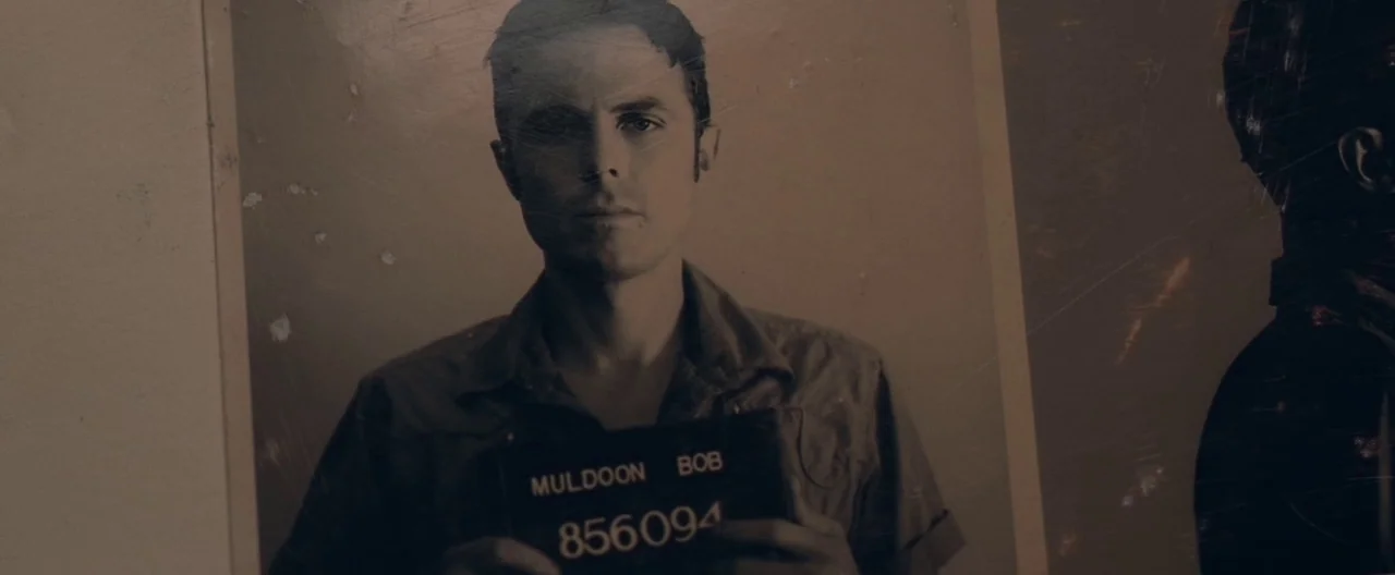 Ain’t Them Bodies Saints (2013) movie still: medium — A close-up of a vintage-style police booking mugshot displayed on a physical surface. The…