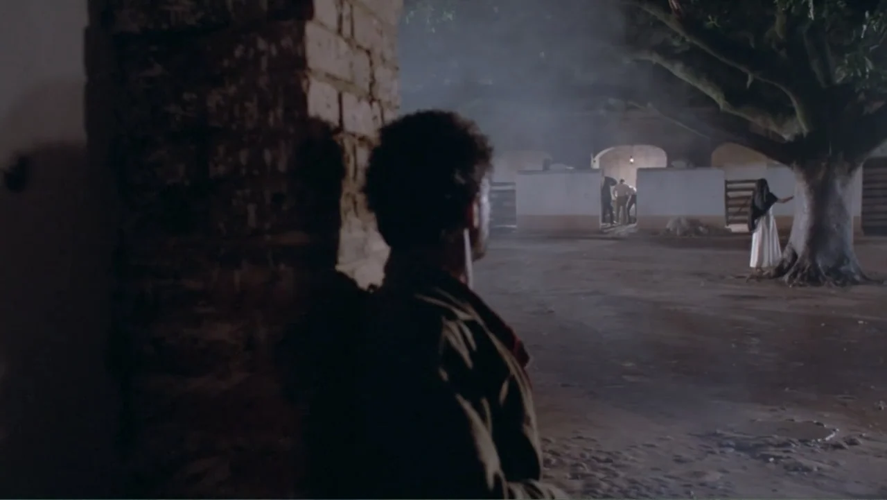 Toña Machetes (1985) movie still: medium — An outdoor courtyard at night features a large central tree and stone walls. A man stands in the…