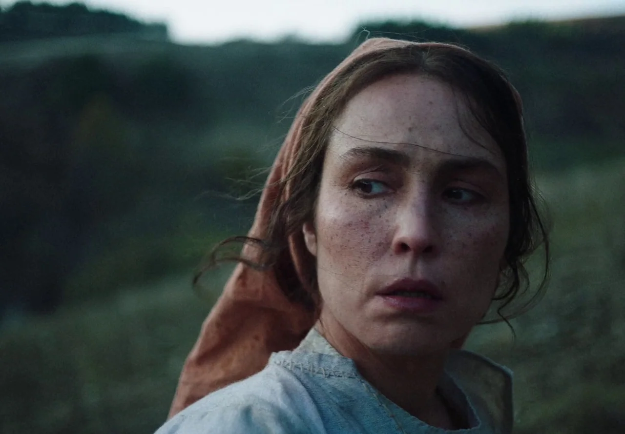 You Won’t Be Alone (2022) movie still: closeup — A woman stands in a dim, grassy landscape during the late evening. She gazes off to the side while…