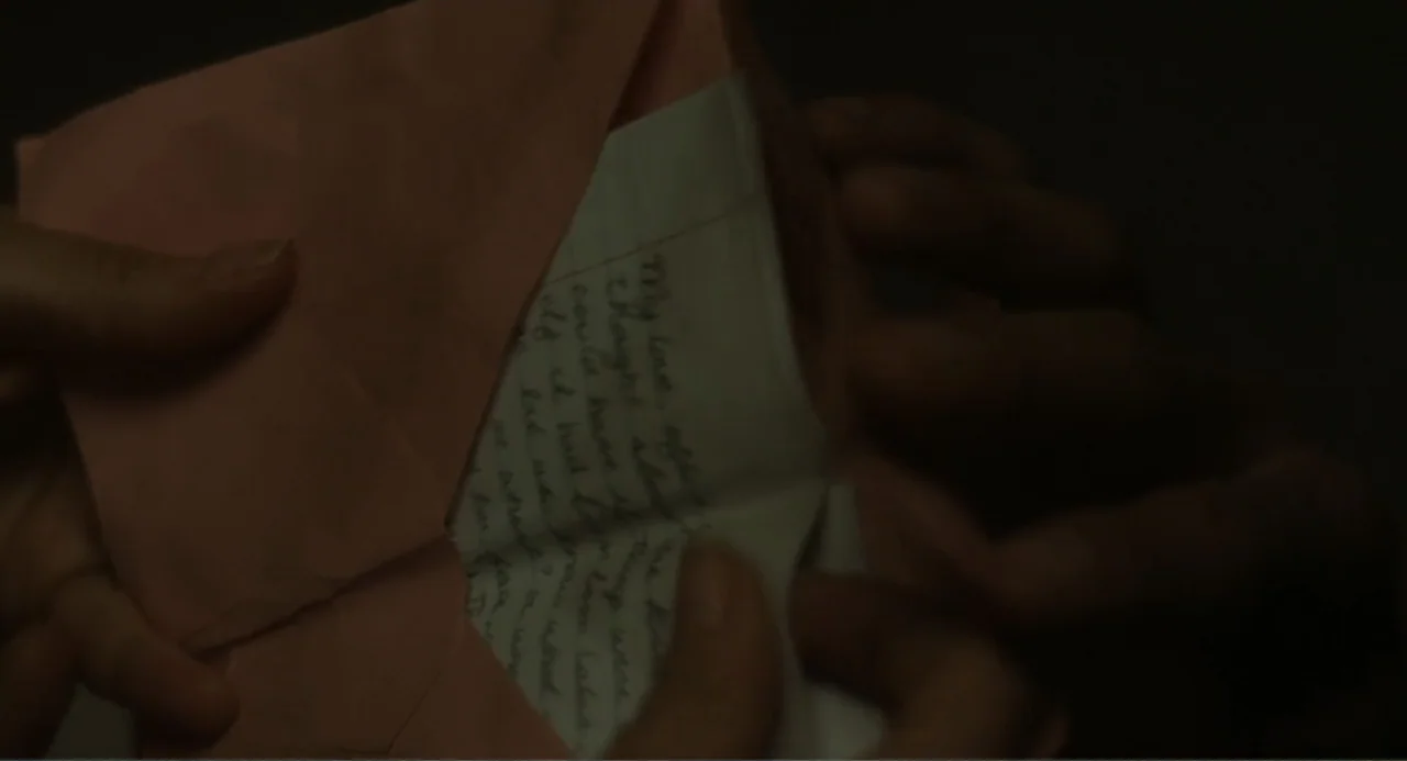 May December (2023) movie still: closeup — A pair of hands holds a folded piece of lined stationery inside a pink envelope. The lighting is low and…