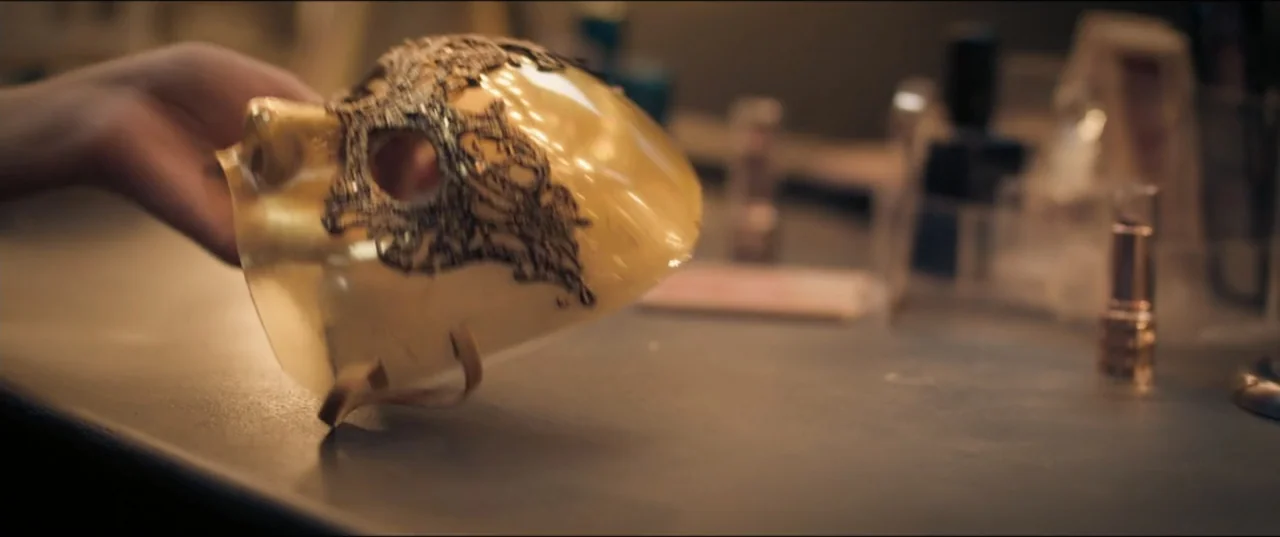 Risqué (2025) movie still: closeup — A vanity surface holds cosmetic items and a gold masquerade mask with black filigree. A hand extends into the…