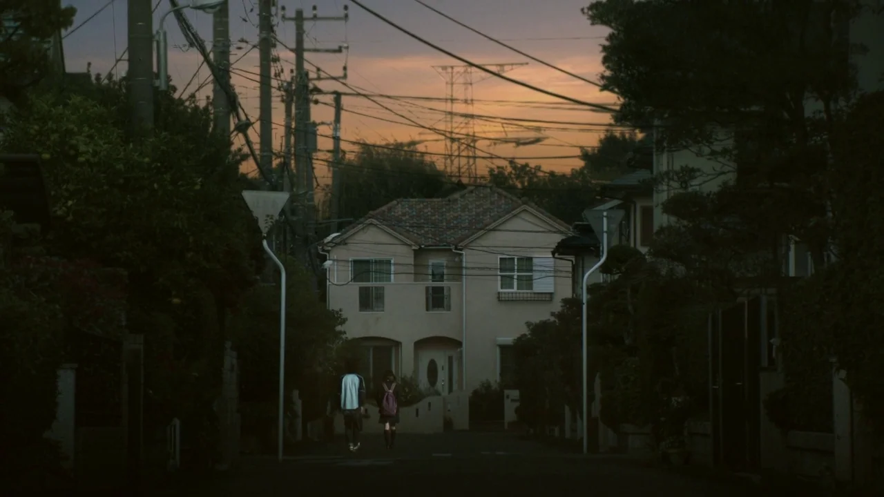 Confessions (2010) movie still: wide — A quiet suburban street in Japan is flanked by lush trees and residential houses. Two teenagers walk away…