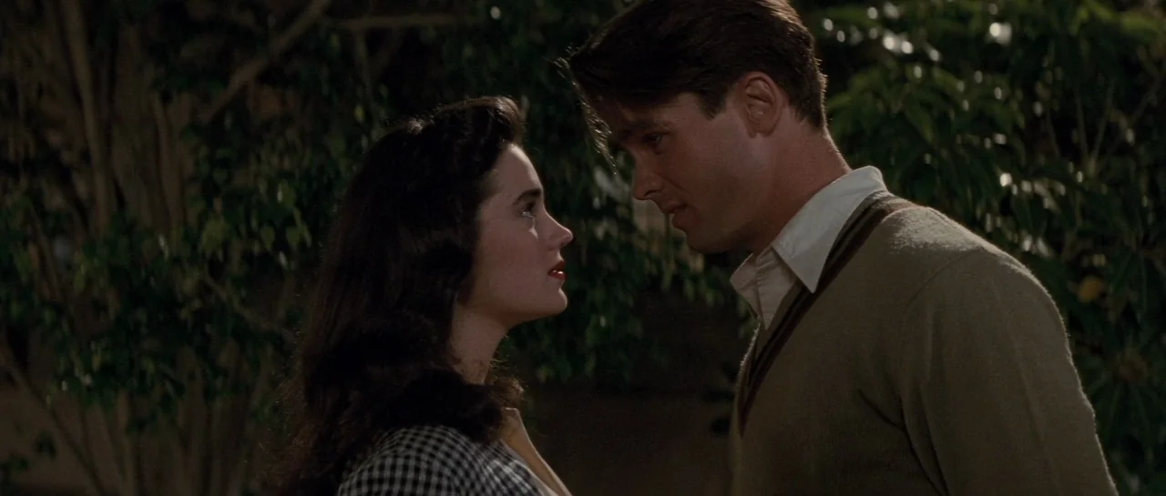 The Rocketeer (1991) movie still: medium — A woman and a man face each other closely in a dimly lit setting. Large leafy bushes surround them in the…