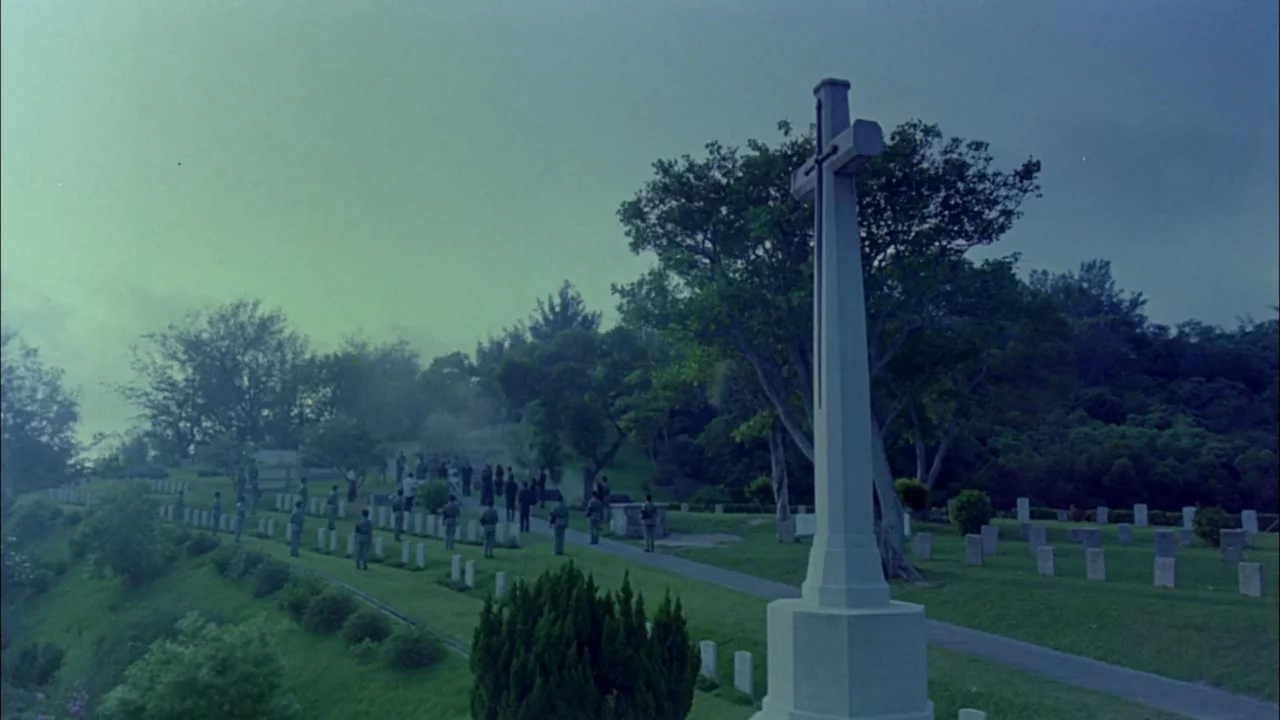 Hard Boiled (1992) movie still: wide — A hillside war cemetery features rows of uniform grave markers and a large stone cross. A small group of…